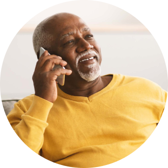 A smiling older Black man with a grey beard wears a yellow sweater while talking on his smartphone